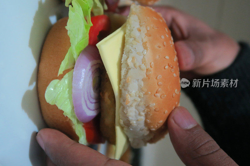 一个男人拿着健康自制的芝士汉堡/汉堡/牛肉汉堡夹在汉堡小面包里，里面有芝麻、融化的奶酪、番茄、红洋葱片和莴苣